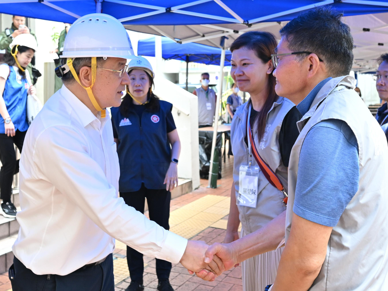 卓永興（左）在大埔廣福社區會堂的裝備穿戴區，與協助上樓居民的地區服務及關愛隊伍義工握手。（政府新聞處）